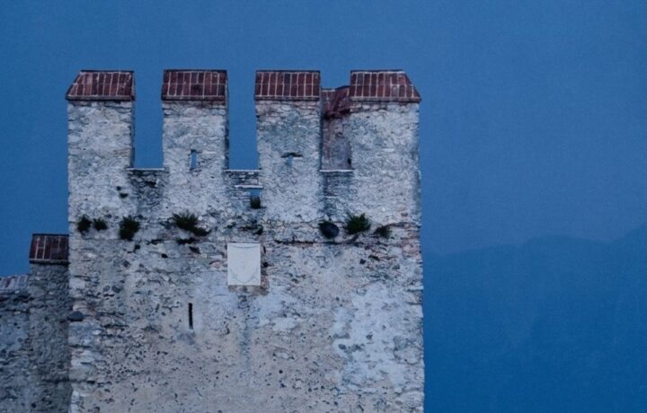 A romantic couple dancing at dusk barefoot on a wooden pier with mooring poles beside the Scaligero Castle tower in Sirmione