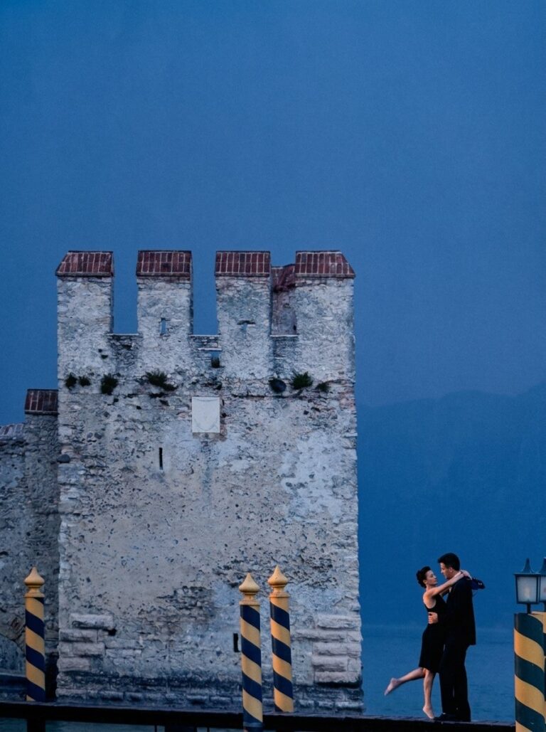 A romantic couple dancing at dusk barefoot on a wooden pier with mooring poles beside the Scaligero Castle tower in Sirmione